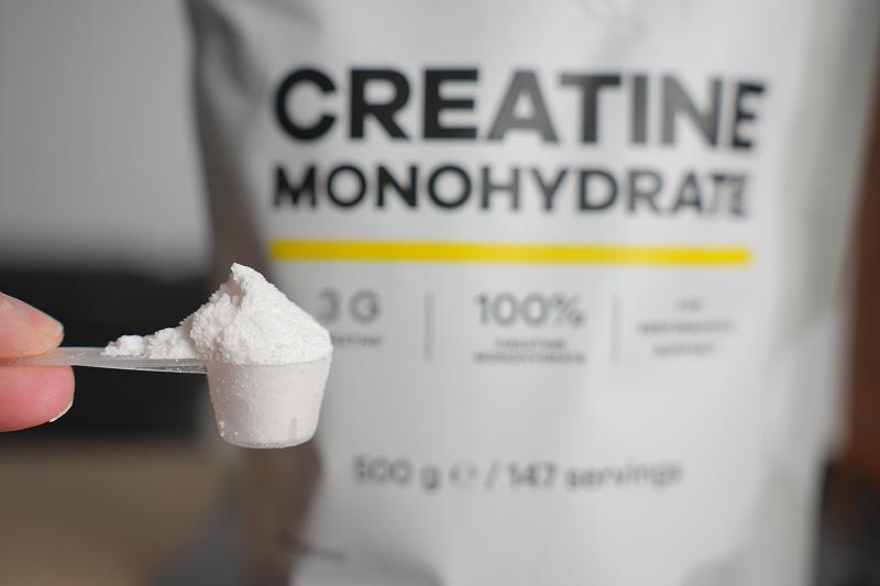 A scoop of white creatine monohydrate powder held in front of a supplement container labeled "Creatine Monohydrate.